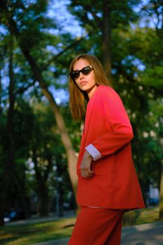 Beautiful blonde woman in a red lady-type pantsuit and black top, watch, ring, sunglasses, with a pendant around her neck is posing sideways while walking alone in the city. The concept of fashion and style. Close-up shot.