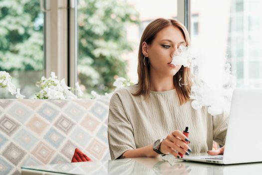 Young Woman Smoking Electronic Cigarette in Restaurant While Working on Laptop