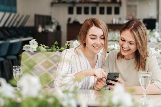 Two Cheerful Women Having Lunch Meeting in Restaurant, Watching on Smartphone and Smiling