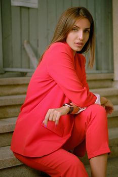 Gorgeous blonde female in a red lady-type pantsuit and black top, watch, ring, with a pendant around her neck is looking at the camera, posing sitting on a stairs while walking alone in the city. The concept of fashion and style. Close-up shot.