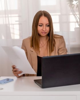 European professional woman sitting with laptop at home office desk, positive woman studying while working on PC. She is wearing a beige jacket and jeans