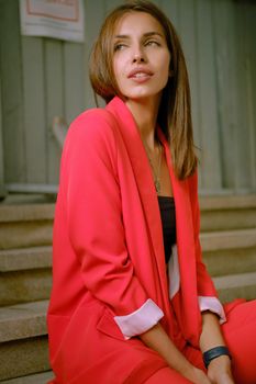 Gorgeous blonde woman in a red lady-type pantsuit and black top, watch, ring, with a pendant around her neck is looking away, posing sitting on a stairs while walking alone in the city. The concept of fashion and style. Close-up shot.