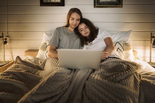 Two beautiful women lying in bed and watching movies on laptop at home