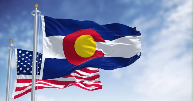 The Colorado state flag waving along with the national flag of the United States of America. In the background there is a clear sky.