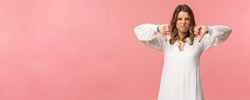 Portrait of picky young blond caucasian woman in white dress over pink background expressing dislike, show thumbs-down and grimacing in aversion, disappointed with bad quality.