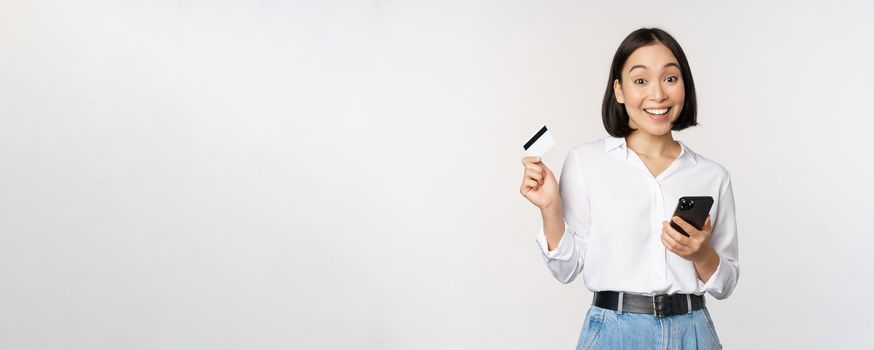 Online shopping concept. Image of young asian modern woman holding credit card and smartphone, buying with smartphone app, paying contactless, standing over white background.