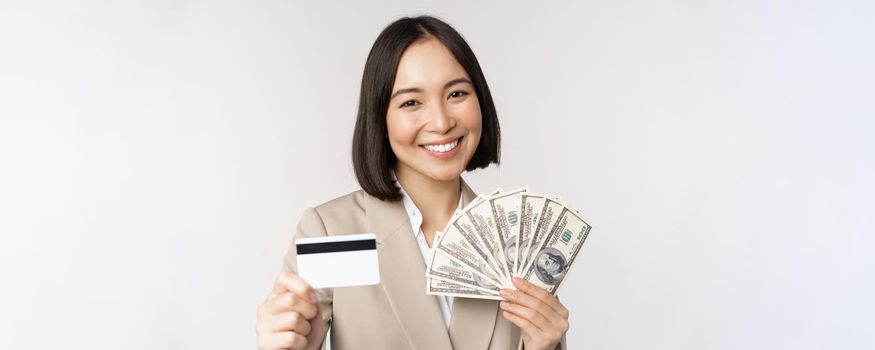 Close up of asian businesswoman, office lady showing credit card and money dollars, standing in suit over white background.