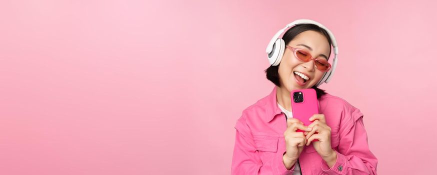 Stylish asian girl in headphones, listening music and taking photos on mobile phone, using smartphone, standing over pink background.