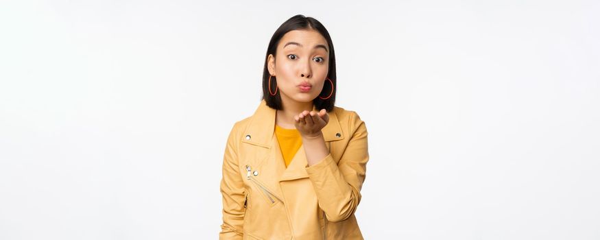 Cute korean stylish girl, sending air kiss, blowing mwah and looking silly romantic at camera, standing over white background. Copy space