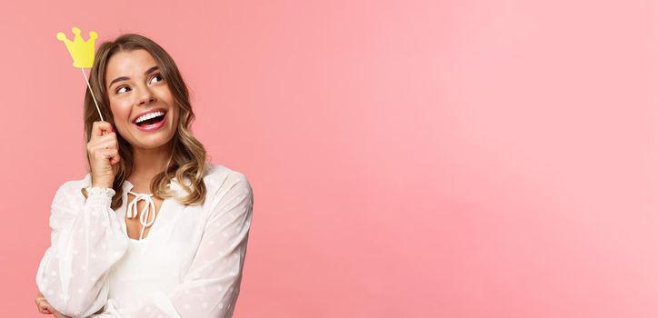 Spring, happiness and celebration concept. Close-up portrait of dreamy beautiful young blond girl imaging something cute and romantic, smiling look up daydreaming with crown on stick, pink background.