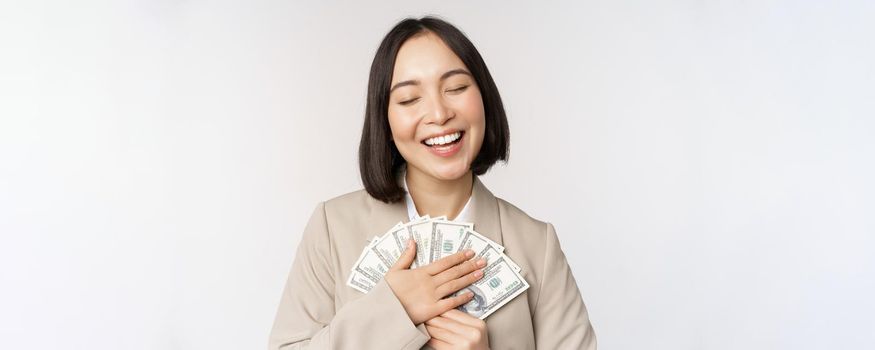 Happy asian businesswoman holding cash, hugging dollars money and smiling, standing over white background in suit.