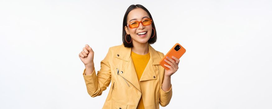 Image of stylish korean girl dancing with smartphone, laughing happy and smiling, standing over white background. Copy space