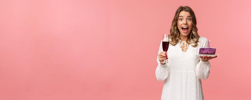 Holidays, spring and party concept. Portrait of fascinated and amused blond girl in white dress look astonished camera, celebrating birthday with b-day cake and glass of wine, pink background.