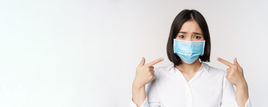 Close up of sad asian girl in medical face mask pointing at her head and looking upset, standing over white background.