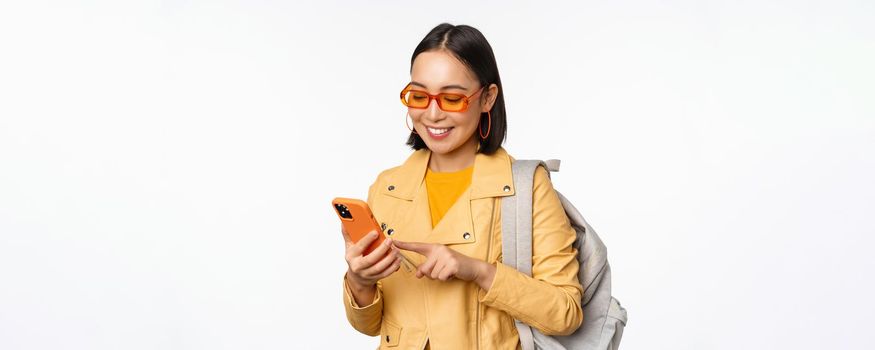 Stylish young asian woman tourist, traveller with backpack and smartphone smiling at camera, posing against white background. Copy space