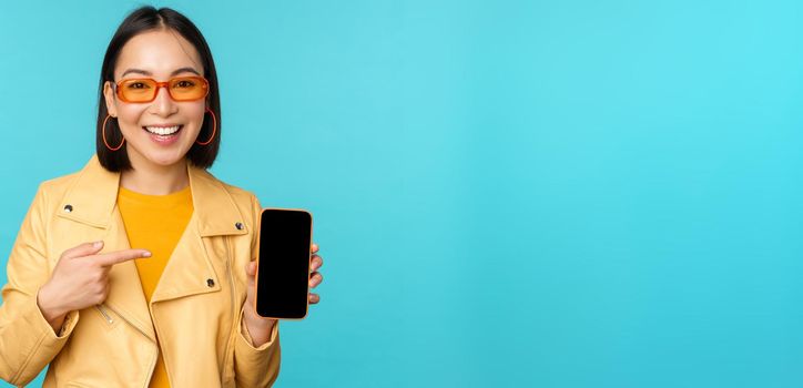Image of young stylish asian female model in trendy sunglasses, showing mobile phone screen, application on smartphone interface, standing over blue background.