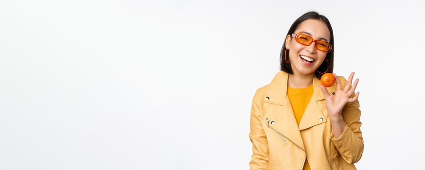 Beautiful asian girl in sunglasses showing tangerine and smiling, looking happy, posing in yellow against studio background.