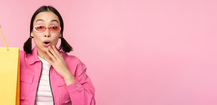 Shopping. Stylish asian girl in sunglasses, showing bag from shop and smiling, recommending sale promo in store, standing over pink background.