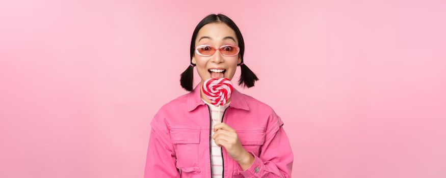 Stylish korean girl licking lolipop, eating candy and smiling, standing in sunglasses against pink background.