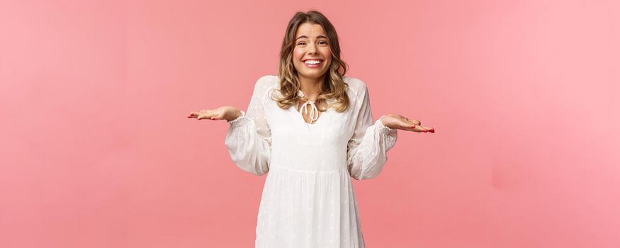 Beauty, fashion and women concept. Sorry dont know, cant say. Cheerful cute silly blond girl 20s, wear white dress, shrugging with hands sideways make awkward smile, apologizing, pink background.