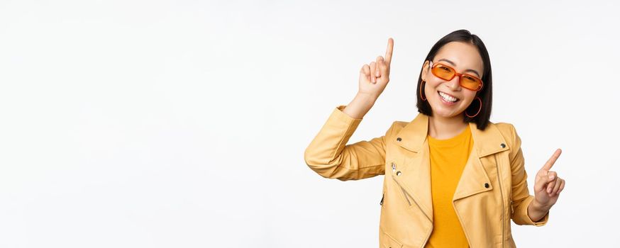 Smiling asian brunette woman in sunglasses, pointing fingers sideways, left and right, showing variants, laughing and dancing, wearing sunglasses, white background.