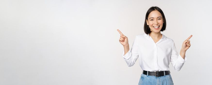 Image of beautiful asian woman pointing fingers left and right, making decision, showing two variants choices, standing over white background.