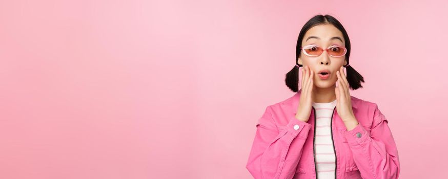 Image of asian girl looking surprised and excited, smiling, amazed reaction to big news, standing over pink background.