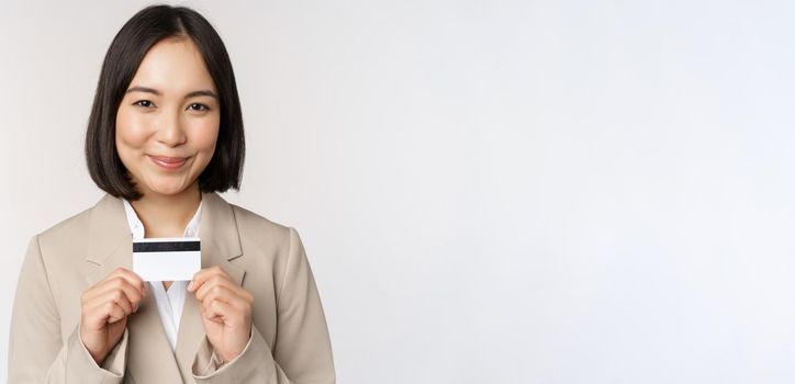 Smiling office clerk, asian corporate woman showing credit card, standing over white background in beige suit. Copy space