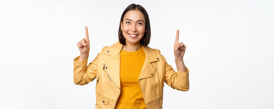 Image of smiling asian brunette woman pointing fingers up, showing advertisement with happy face, posing against white background.