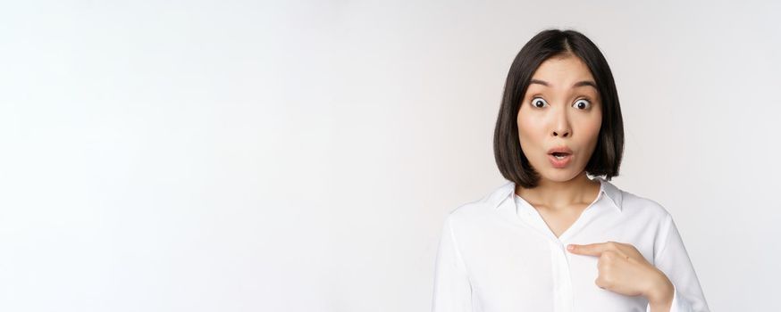 Close up portrait of young asian woman pointing finger at herself with disbelief, surprised that she was named, standing over white background.