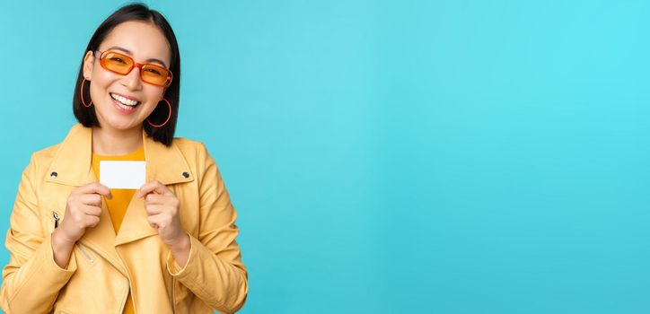 Stylish young asian woman in sunglasses, showing credit card and smiling, recommending bank, contactless payment or discounts in store, standing over blue background.