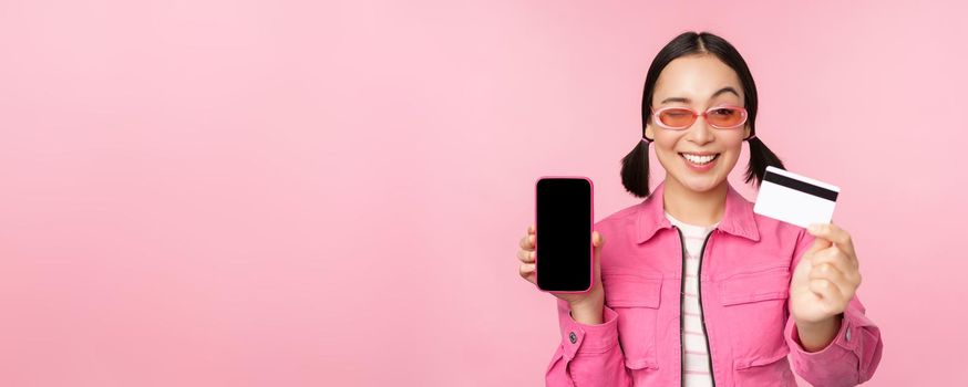 Image of smiling korean woman showing credit card and mobile phone screen, smartphone application interface, paying online, shopping contactless, standing over pink background.