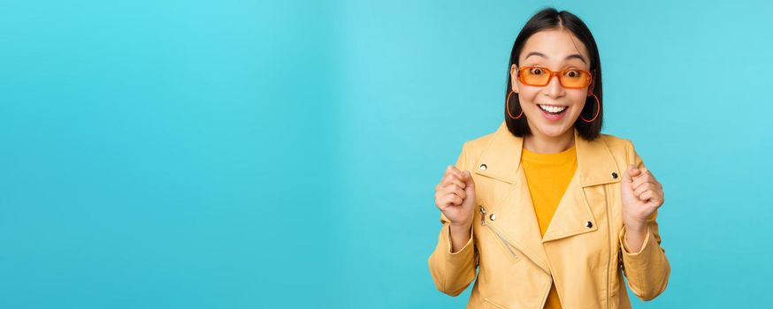 Image of enthusiastic young asian woman celebrating, triumphing, looking surprised and happy, clapping hands satisfied, standing over blue background.