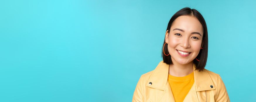 Close up portrait of smiling beautiful asian woman with white teeth, looking happy at camera, posing in yellow jacket over blue studio background.