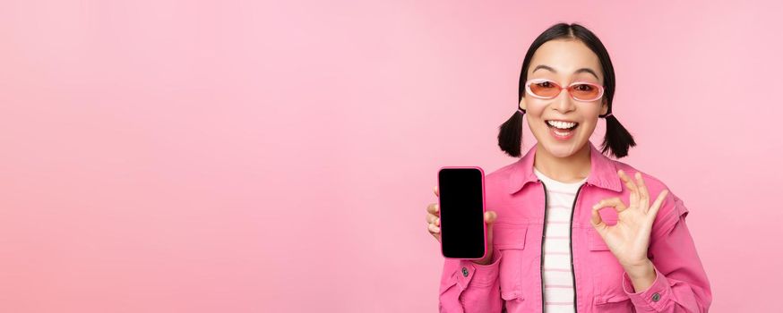 Enthusiastic young asian woman showing okay, ok sign, smiling pleased, mobile phone screen, smartphone application, standing over pink background.