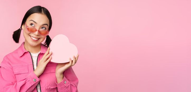 Image of stylish asian girlfriend in sunglasses, guessing whats inside gift box, heart-shaped present, standing over pink background.