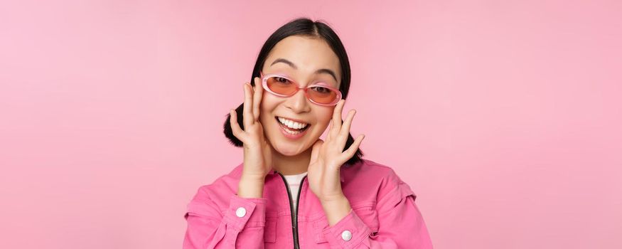 Cute modern japanese girl in sunglasses, smiling and looking happy, posing against pink background in stylish clothing.