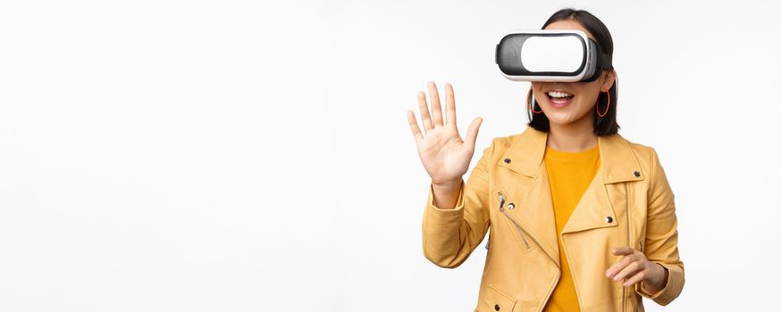 Happy asian girl in VR headset, saying hello in virtual reality chat, using special glasses, standing over white background, waving hand, saying hello.