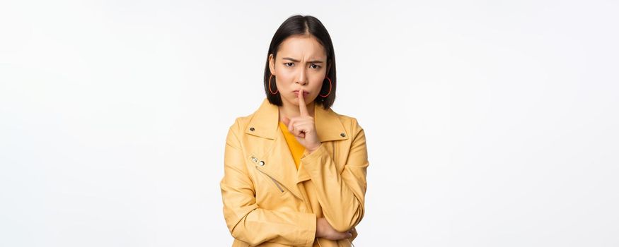 Portrait of angry korean girl shushing, woman frowning and tell to be quiet, showing hush, shh gesture, press finger to lips, taboo sign, standing over white background.