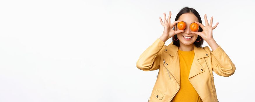 Funny asian girl holding tangerine on eyes and smiling, making playful grimaces, standing over white background. Copy space