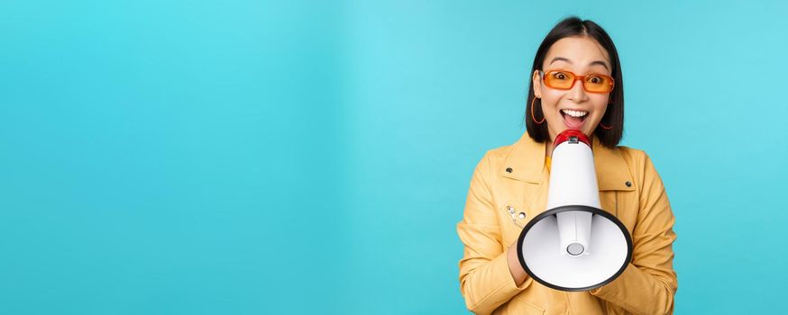 Stylish asian girl making announcement in megaphone, shouting with speakerphone and smiling, inviting people, recruiting, standing over blue background.