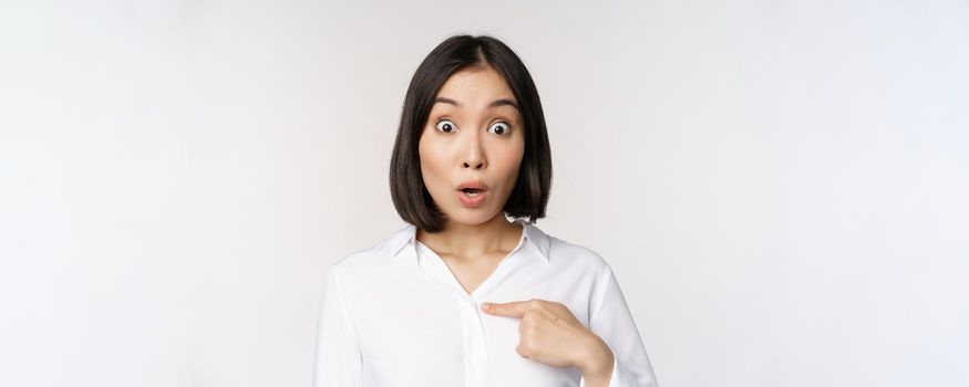 Close up portrait of young asian woman pointing finger at herself with disbelief, surprised that she was named, standing over white background.