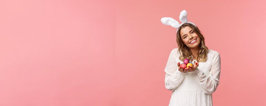 Holidays, spring and party concept. Portrait tender, romantic blond girl in white dress and rabbit ears, tilt head cute, smiling happy as holding painted eggs, celebrating Easter, pink background.
