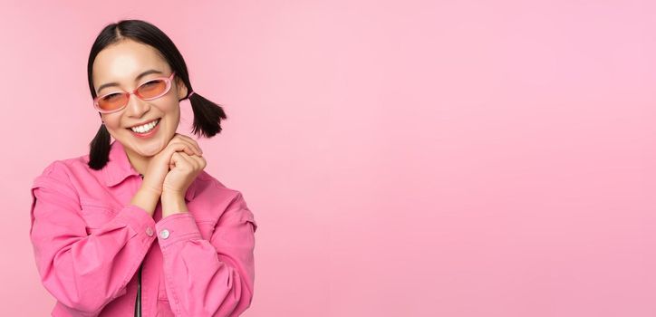 Close up portrait of stylish asian woman in sunglasses, smiling, looking flattered, laughing coquettish, standing over pink background. Copy space