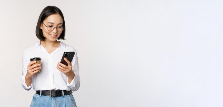 Image of modern asian woman looking at mobile phone, drinking takeaway coffee, wearing glasses, using smartphone app, standing over white background.