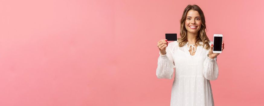 Portrait of satisfied cute blond girl in white dress, showing you her credit card and mobile phone application, online shopping app or bank website, smiling camera, standing pink background.