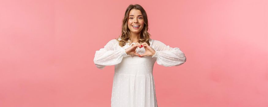 Beauty, fashion and women concept. Romantic passionate blond european female in white cute dress, make heart sign near chest and smiling at camera excited, show sympathy.