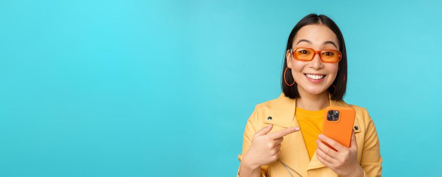Enthusiastic asian girl in stylish sunglasses, pointing finger at smartphone, showing mobile phone, standing over blue background.