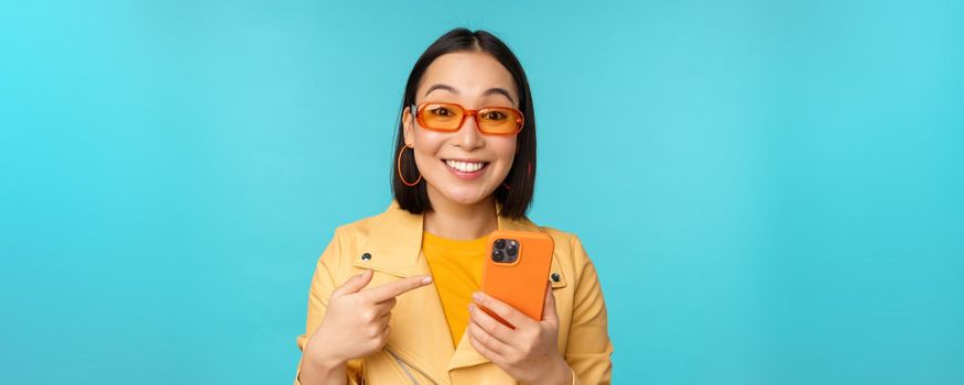 Enthusiastic asian girl in stylish sunglasses, pointing finger at smartphone, showing mobile phone, standing over blue background.