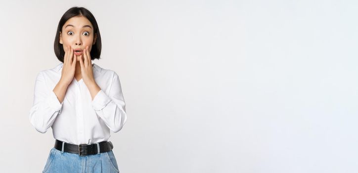 Portrait of surprised young office woman, asian businesswoman gasping amazed, saying wow, standing impressed of news against white background.